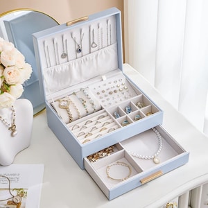 May include: A light blue jewelry box with a hinged lid and a pull-out drawer. The interior features hooks for necklaces, a padded section for rings, multiple compartments for earrings, and a velvet-lined tray for bracelets and necklaces.
