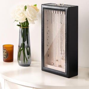 May include: A rectangular black jewelry display case with a clear glass front, featuring a beige interior lining with multiple silver-toned necklaces hanging vertically. The case has a silver metal latch at the top and a stitched leather-like frame.