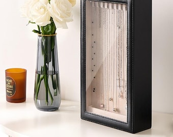 May include: A rectangular black jewelry display case with a clear glass front and a silver latch at the top. The interior features a light-colored fabric backing with multiple silver-toned necklaces hanging vertically from hooks inside the case.