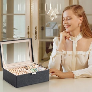 May include: A black jewelry box with a mirrored lid is open, revealing various rings and bracelets. The box sits on a white surface. A woman is seated next to the box, resting her chin on her hand.
