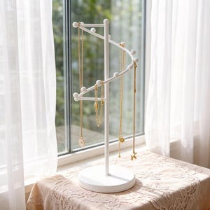 May include: A white metal jewelry display stand with a spiral design. Several gold necklaces with various pendants hang from the stand. The stand is on a round base and sits on a table with a lace tablecloth, near a window with white curtains.