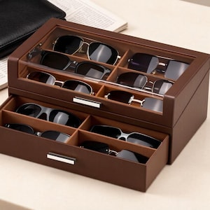May include: A brown leather-textured storage box for eyewear featuring a glass-topped upper compartment and a pull-out drawer. The interior is divided into multiple sections, each containing a pair of dark-tinted sunglasses with metallic frames.
