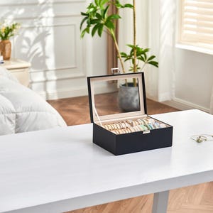 May include: A black jewelry box with a clear glass lid, open to display various bracelets and rings. The box is on a white table, with a plant and window in the background. The jewelry is gold, silver, and colorful.