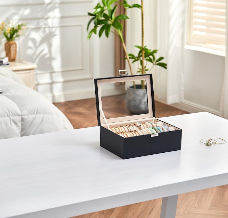 May include: A black jewelry box with a clear glass lid, open to display various bracelets and rings. The box is on a white table, with a plant and window in the background. The jewelry is gold, silver, and colorful.