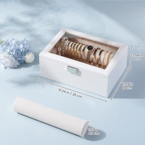 May include: White jewelry box with a clear top, holding various gold bracelets and a watch. The box is 10.24 inches long, 7.09 inches wide, and 3.54 inches tall. A white, cylindrical jewelry display is in the foreground, 1.93 inches in diameter.
