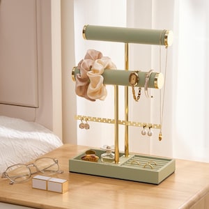 May include: A sage green and gold jewelry organizer stand featuring two horizontal bars for bracelets and necklaces, a perforated metal strip for earrings, and a rectangular base with a tray and ring slots. The stand holds a beige scrunchie, a gold watch, a bracelet, and a long necklace.