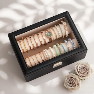 May include: A black jewelry box with a glass top, filled with various bracelets and a watch. The bracelets are gold and rose gold, with some featuring beads and shells. A brown leather watch with a green face is also present. Two light-colored roses are in the foreground.