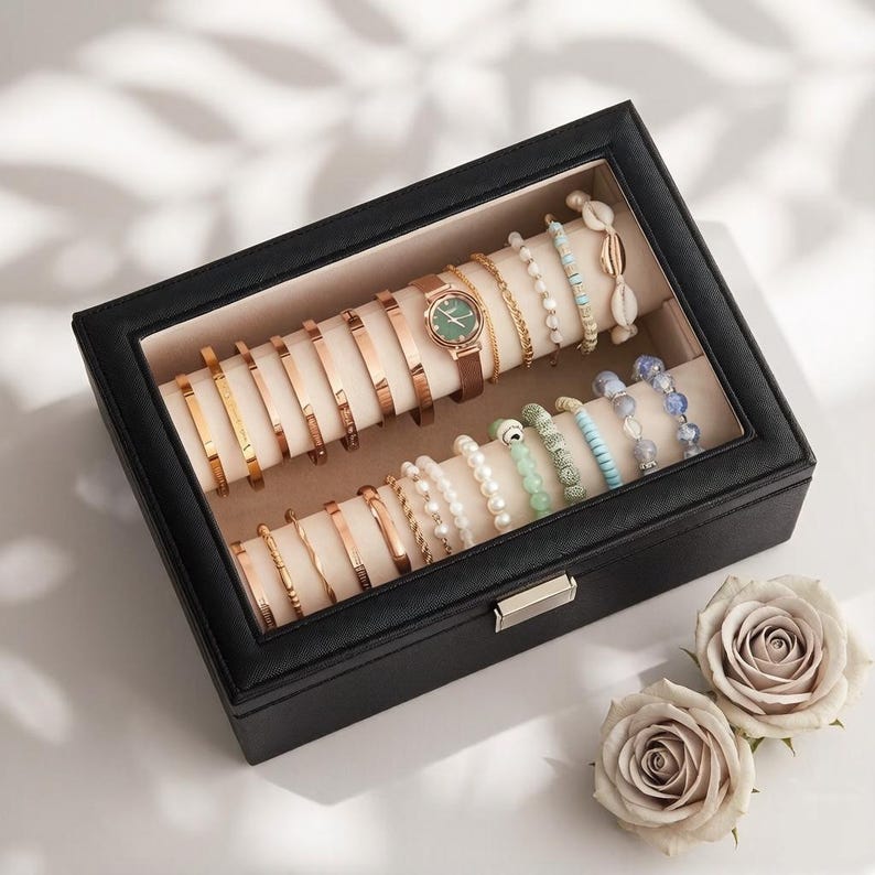 May include: A black jewelry box with a glass top, filled with various bracelets and a watch. The bracelets are gold and rose gold, with some featuring beads and shells. A brown leather watch with a green face is also present. Two light-colored roses are in the foreground.