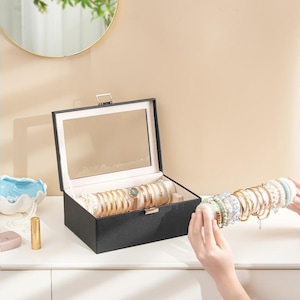 May include: A black jewelry box with a mirrored lid, open to display gold bracelets and a watch. A hand holds a rod of bracelets, showcasing various colors and designs. The box sits on a white surface.