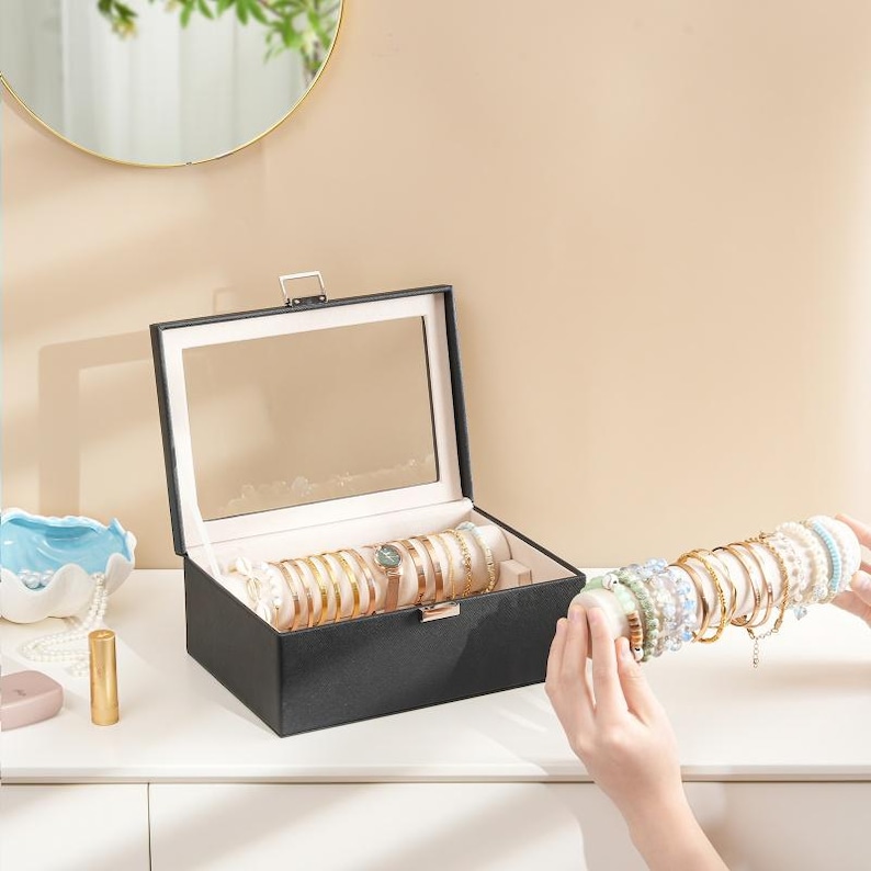 May include: A black jewelry box with a mirrored lid, open to display gold bracelets and a watch. A hand holds a rod of bracelets, showcasing various colors and designs. The box sits on a white surface.