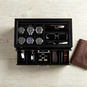 May include: A black wooden watch and accessories organizer with a glass top. The top section displays several watches and sunglasses. The bottom drawer holds more accessories, including rings, bracelets, and a watch. A brown leather wallet is on the side.