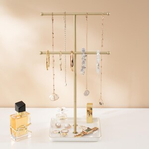 May include: A gold-toned jewelry display stand with multiple necklaces, bracelets, and earrings. The stand has a rectangular base with a white surface and gold trim. Perfume bottles, a watch, and rings are also displayed.