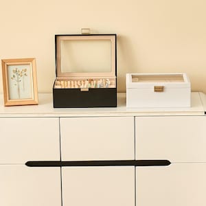 May include: A wooden framed picture and two jewelry boxes sit atop a white cabinet. The black jewelry box is open, revealing jewelry. The white box is closed. The picture frame is light brown.