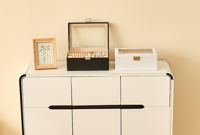 May include: A wooden framed picture and two jewelry boxes sit atop a white cabinet. The black jewelry box is open, revealing jewelry. The white box is closed. The picture frame is light brown.