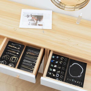 May include: Open drawers of a white jewelry organizer filled with various necklaces, bracelets, and rings. The jewelry is displayed on black velvet trays. The organizer sits on a light wood surface with a mirror and a bottle.