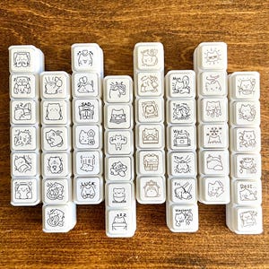 May include: A collection of white square stamps with black line art illustrations. The stamps feature various cat-themed designs and text, including emotions, daily activities, and days of the week. The stamps are arranged on a wooden surface.