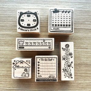 Op de afbeelding: Een verzameling houten stempels met kat-thema ontwerpen. Inclusief een klok met "Meow's Clock", een kalender, een weekplanner, een huis met een datum, een takenlijst en een verticale stempel met nummers en katten.