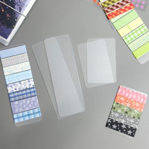 Peut inclure: Ensemble de protège-cartes rectangulaires transparents et de bandes de washi tape décoratives. Le washi tape présente divers motifs, dont des carreaux, des étoiles et des motifs géométriques dans des tons de bleu, vert, rose et noir. Les articles sont disposés sur une surface grise.