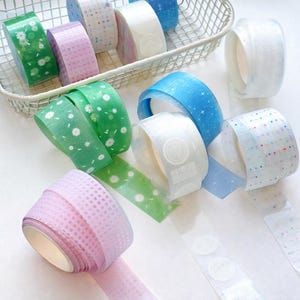 May include: A selection of decorative washi tape rolls in various colours and patterns. The tape designs include floral, polka dots, and geometric patterns. Some rolls are green, pink, blue, and white. The tape is displayed in a wire basket and scattered on a white surface.