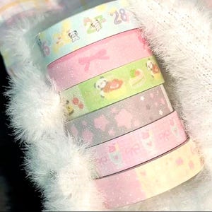May include: A collection of pastel-coloured washi tapes. The tapes display patterns such as bows, stars, panda bears, and cakes. Some tapes have glitter and the number "28".