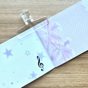 May include: A clear plastic hair clip securing a light blue paper sheet featuring a dark treble clef symbol, scattered light blue stars, and a section with a light purple lily floral pattern and a subtle white polka dot background design.