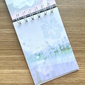 May include: A spiral-bound notebook with white metal rings featuring a light blue and white page design. The paper displays a subtle, repeating hibiscus flower pattern in a soft, muted blue tone, with a blurred, abstract gradient effect across the center.