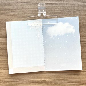 May include: An open notebook held by a clear plastic clip. The left page features a light blue grid pattern and a white umbrella graphic with the word dream. The right page shows a white cloud against a light blue sky with white stars.