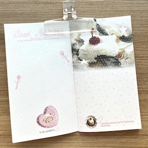 May include: A rectangular paper notepad held by a clear plastic clip, featuring a light pink polka dot pattern, illustrations of cutlery, a heart-shaped plate with a cake roll, a dessert image with a cherry, and text including Sweet Archive.