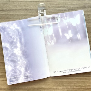 May include: An open notebook held by a clear plastic clip, featuring light purple pages with white floral and butterfly motifs. The left page displays English text, while the right page includes Japanese and Korean script at the top and bottom.