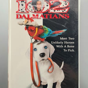 May include: A VHS tape for Disney's 102 Dalmatians. The cover features a white puppy with blue eyes and a red and orange leash, a colorful parrot, and the film's title in red. The background is split black and white.