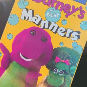 May include: A VHS tape from the Barney & Friends Collection, titled "Barney's Manners." The cover features Barney the dinosaur and a green dinosaur character. The background is yellow with blue bubbles. Includes a free parent/child activity guide.