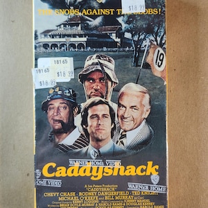 Może przedstawiać: Vintage kaseta VHS z filmem "Caddyshack" z kolażem twarzy aktorów. Okładka zawiera logo Warner Home Video i tytuł filmu w kolorze żółtym. Naklejki z cenami są widoczne w lewym górnym rogu.