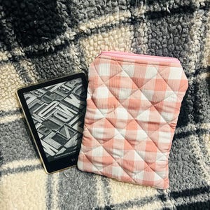 May include: An e-reader with a black screen displaying text, next to a pink and white checked quilted case with a zip. The case is designed to protect the e-reader. The background is a textured, patterned fabric.
