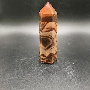 Op de afbeelding: Een gepolijste, zeshoekige stenen obelisk. De bovenkant is rood-oranje, overgaand in bruine en bruine banden. De steen heeft een wervelpatroon en de basis is donkerbruin. Een decoratief object.