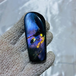 Puede incluir: Una piedra preciosa de labradorita pulida y rectangular. La piedra muestra un vibrante juego de colores, con azules iridiscentes, púrpuras y destellos amarillos. La gema se sostiene en un guante gris.