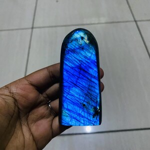 May include: A polished labradorite stone with a rounded top, featuring a vibrant, iridescent blue flash across its surface. The stone displays natural internal fractures and dark inclusions, creating a textured appearance within the deep blue mineral.
