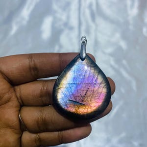 May include: A teardrop-shaped labradorite pendant with a silver bail. The stone exhibits a vibrant play of colour, including blues, purples, and pinks. The pendant is held in a hand, showcasing the polished surface and unique patterns.
