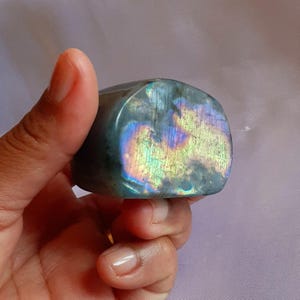 May include: A polished labradorite stone with a rounded shape. The stone displays iridescent flashes of blue, purple, and gold across a gray base. The stone is held in a hand.