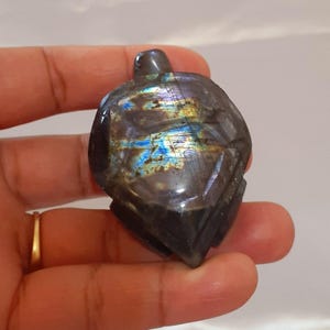 Peut inclure: Une pierre précieuse en labradorite sculptée en forme de tortue. La pierre est principalement gris foncé avec des reflets irisés bleus, verts et jaunes. La tortue a des détails sculptés sur sa carapace et sa tête, tenue dans une main.