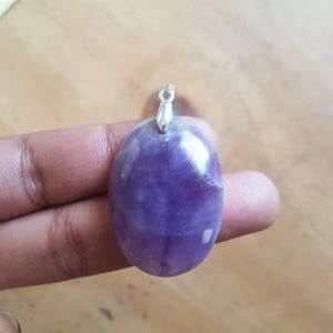 Puede incluir: Un colgante de amatista ovalada pulida, con una argolla plateada. La piedra preciosa presenta un color púrpura intenso con vetas más claras. El colgante está diseñado para ser usado como collar.