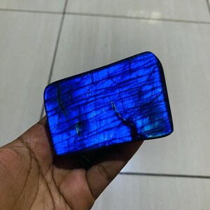 May include: A rectangular polished labradorite stone displaying a vibrant, iridescent electric blue flash across its surface. The stone features a network of dark, thin, linear inclusions and natural fractures that contrast with the bright blue color.