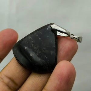 Può includere: Un ciondolo in pietra preziosa nera lucida, di forma triangolare con bordi arrotondati, con una cauzione color argento. La superficie è liscia e riflettente. La cauzione è fissata alla parte superiore della pietra con un piccolo anello per una catena.