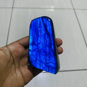 Peut inclure: Une pierre de labradorite polie présentant un éclat bleu électrique vibrant sur toute sa surface. La pierre possède un bord supérieur lisse et arrondi ainsi qu'une base plate, avec des fissures sombres et complexes en forme de toile.