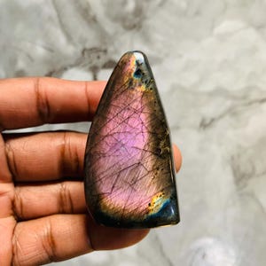 May include: A polished labradorite gemstone with a triangular shape, featuring a prominent iridescent pink flash across the centre, dark grey and black inclusions, and subtle hints of blue and gold along the edges with a smooth, reflective surface.