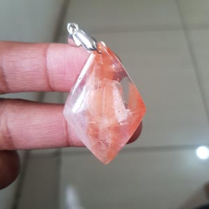 Può includere: Un ciondolo trasparente a forma di diamante con marmorizzazione interna arancione e bianca. Il ciondolo ha un'attaccatura color argento per il fissaggio a una catena. La gemma è tenuta dalle dita di una persona.
