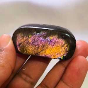 May include: A polished, rectangular labradorite gemstone. The stone has a dark base color with iridescent flashes of purple, yellow, and orange. The gemstone is held in a hand, showcasing its smooth, rounded edges and natural beauty.
