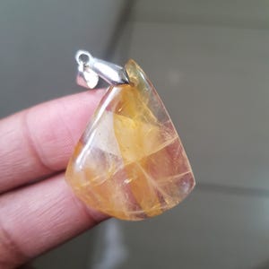 Può includere: Un ciondolo triangolare con una cauzione d'argento. Il ciondolo è realizzato in una pietra preziosa giallo-oro traslucida con fratture interne. La pietra ha una superficie lucida e un aspetto sfaccettato.