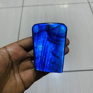 Puede incluir: Una piedra de labradorita pulida que presenta un vibrante destello azul eléctrico en su superficie. El mineral muestra una forma rectangular lisa con esquinas redondeadas y fracturas internas visibles que captan la luz, creando un brillo profundo.