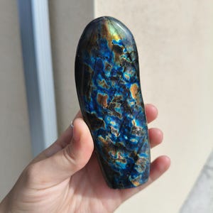 May include: A polished, elongated labradorite gemstone. The stone displays a vibrant play of color, with iridescent blues, greens, and hints of gold. The surface has natural patterns and textures, making each piece unique.