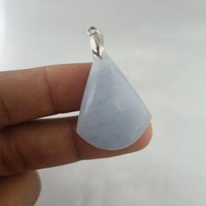 Peut inclure: Un pendentif triangulaire bleu clair avec une bélière argentée. Le pendentif est tenu dans une main, mettant en valeur sa surface lisse et polie. La pierre précieuse est translucide.